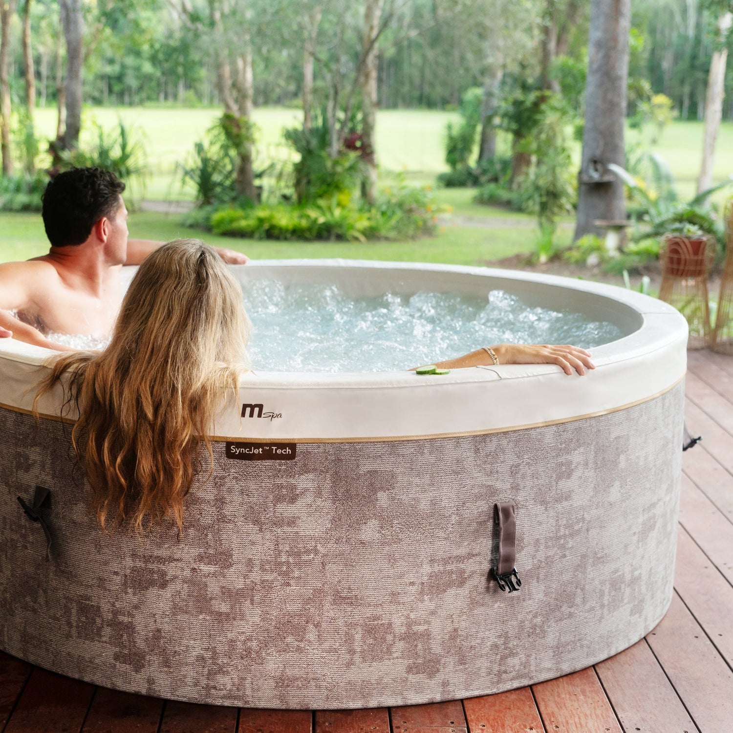 Image of a couple relaxing while having a spa in a hot tub jacuzzi positioned on a wooden deck looking out at a beautiful scene of green grass and trees.