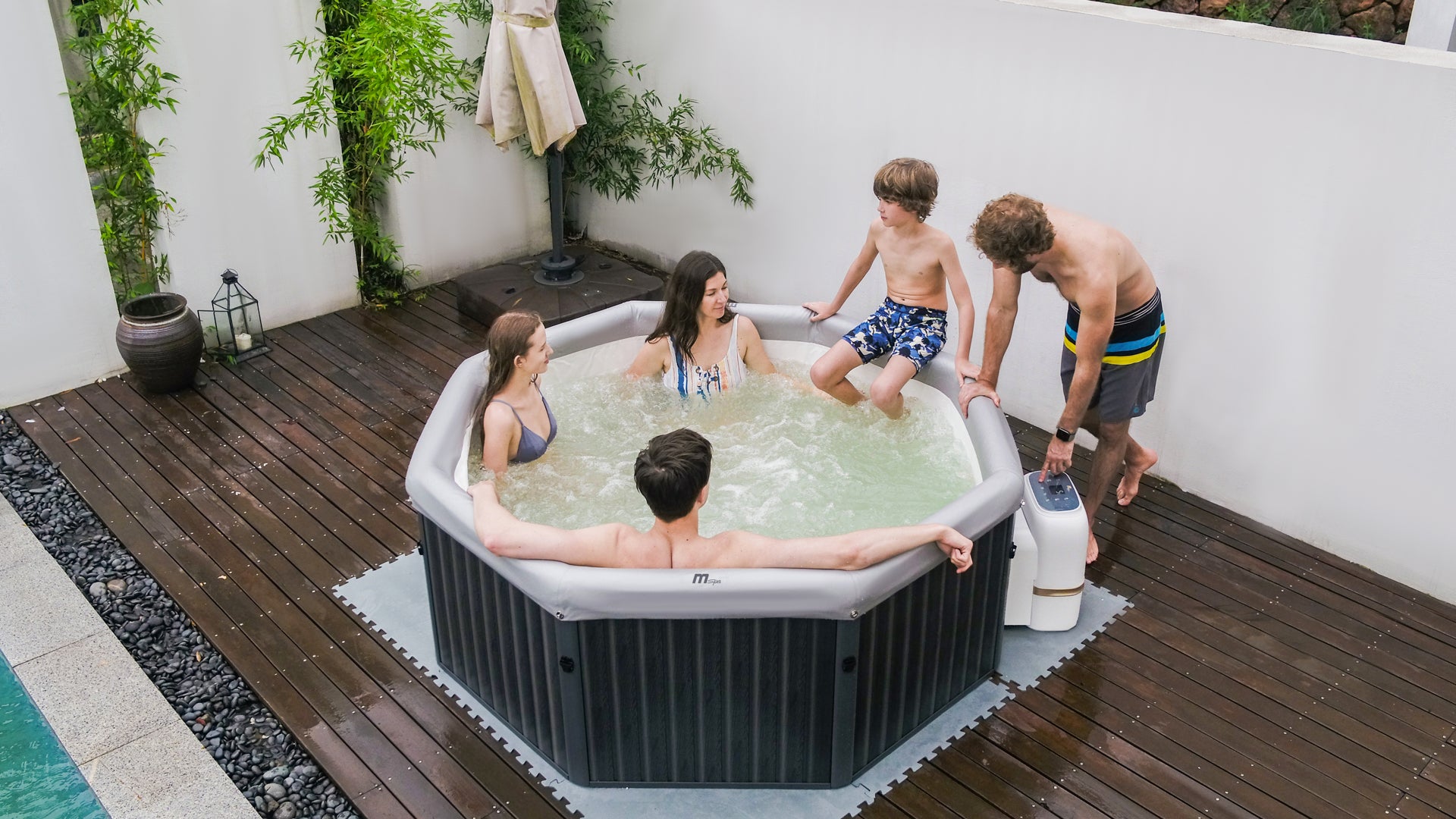 Family enjoying a spa in inflatable portable black hot tub jacuzzi on a wooden deck with plants next to a pool