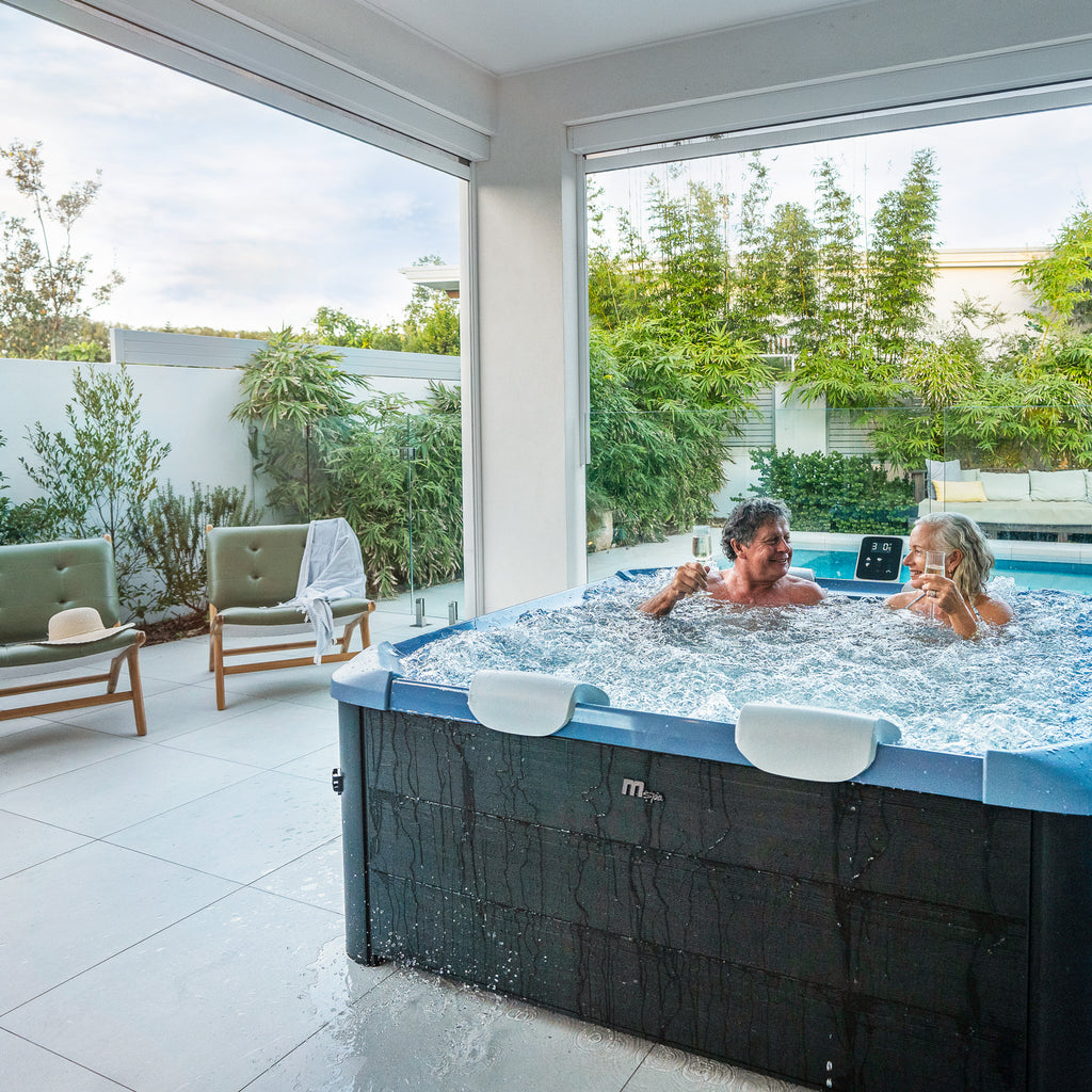 A happy middle aged couple enjoying a spa in a square framed hot tub jacuzzi with black wood like exterior and a sapphire blue interior, situated in a covered outdoor area with outdoor furniture and a pool surrounded by green plants in the background.