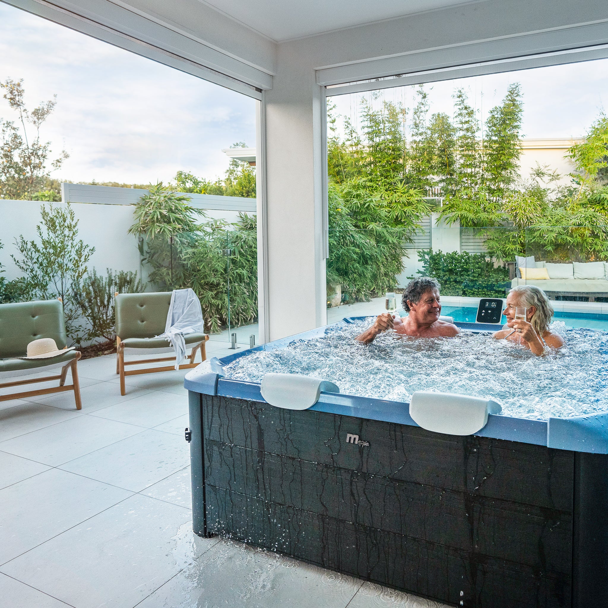 A happy middle aged couple enjoying a spa in a square framed hot tub jacuzzi with black wood like exterior and a sapphire blue interior, situated in a covered outdoor area with outdoor furniture and a pool surrounded by green plants in the background.