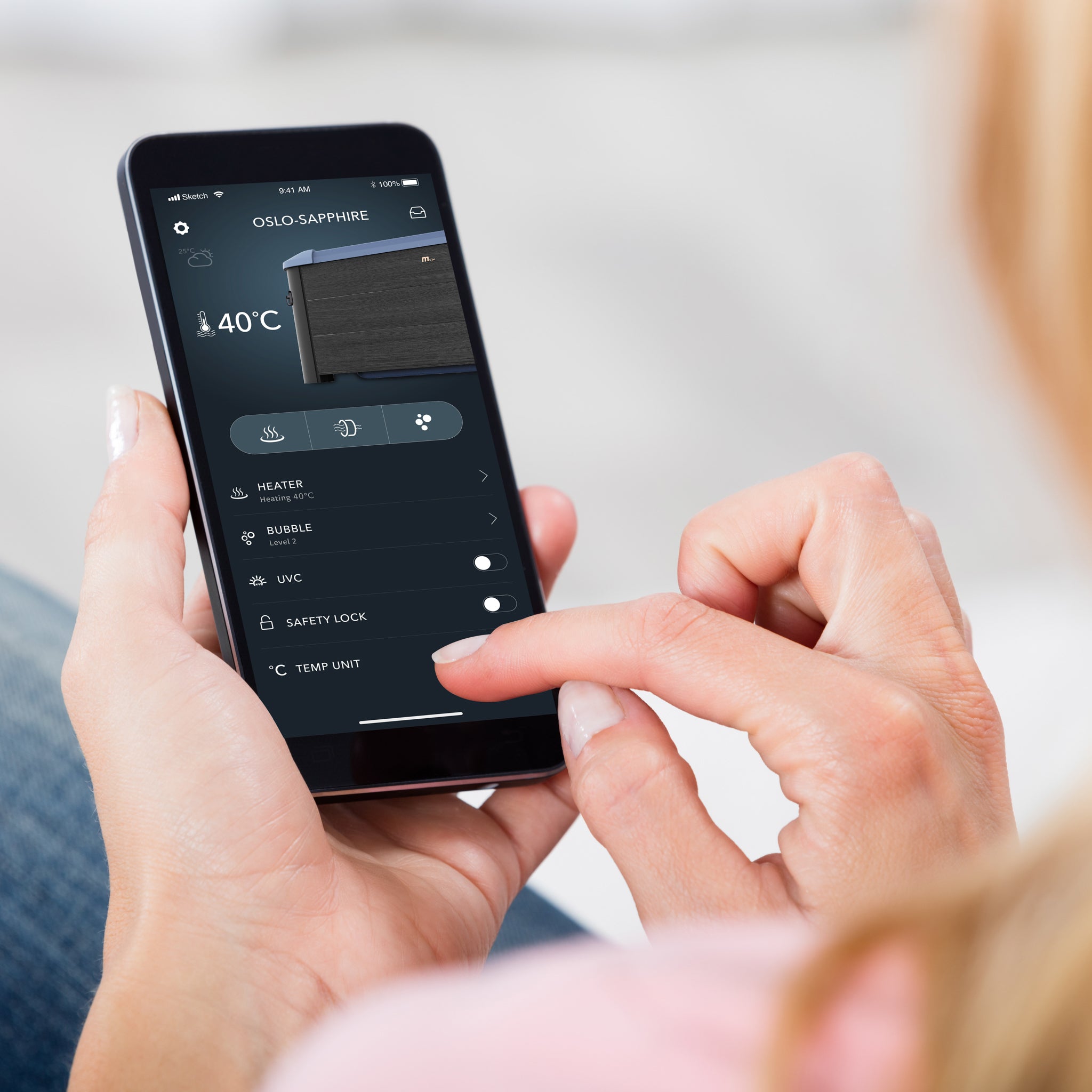 A close up of a woman's hands holding a smartphone with the MSpa App open on her screen, showing the temperature, hydro jet massage and UVC control features of the Oslo Sapphire portable black wood like square framed spa hot tub jacuzzi.