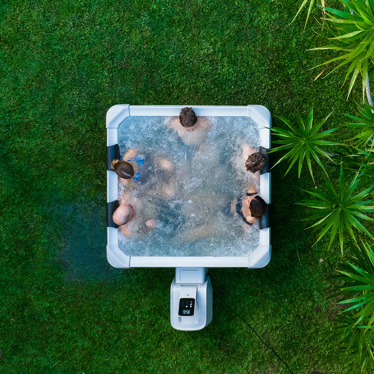 Top-down view of five people relaxing in a square framed hot tub jacuzzi spa surrounded by green grass and plants.