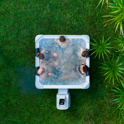 Top-down view of five people relaxing in a square framed hot tub jacuzzi spa surrounded by green grass and plants.