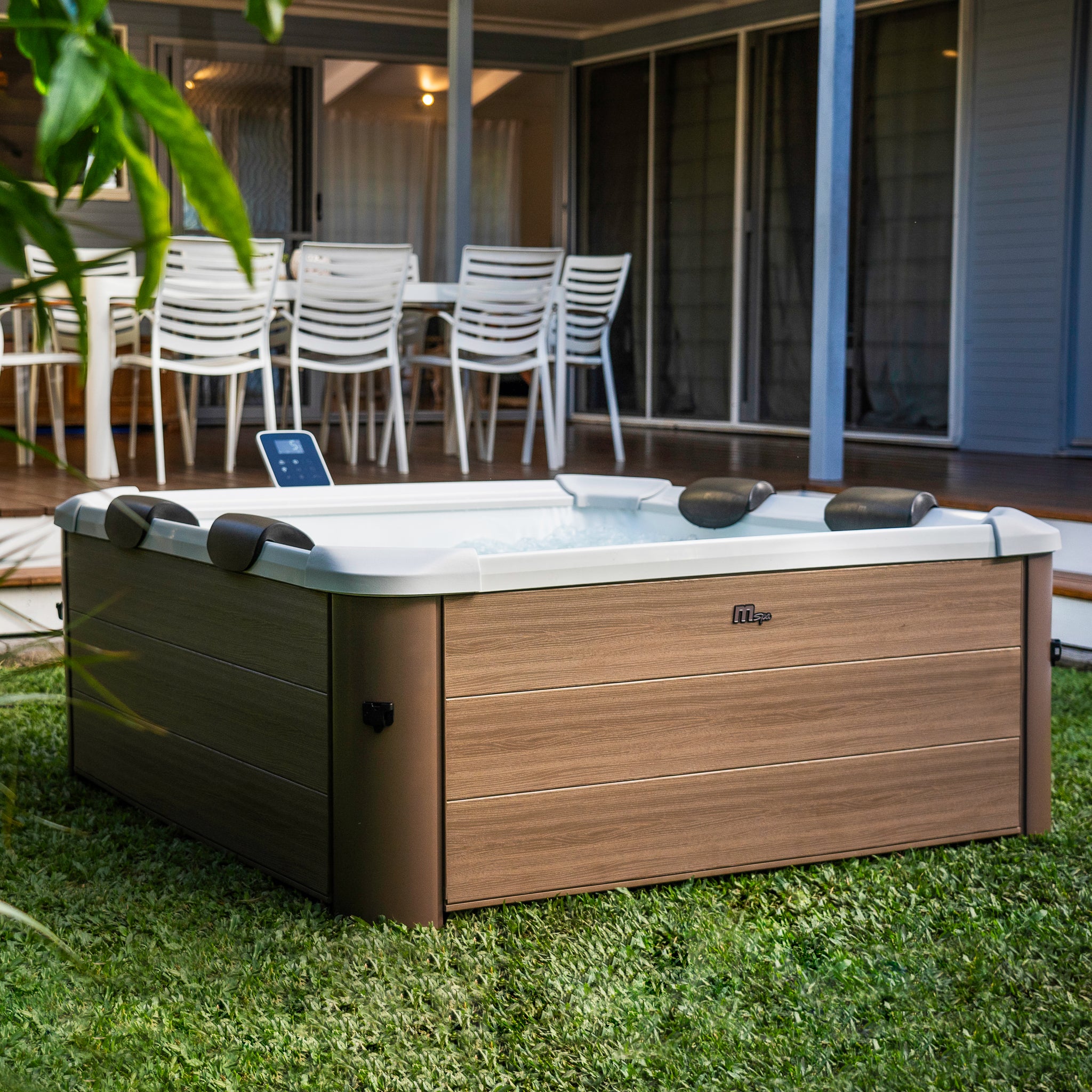 Portable hot tub jacuzzi spa with square wood-like brown frame on a grassy garden area with a patio and chairs in the background