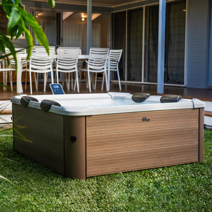 Portable hot tub jacuzzi spa with square wood-like brown frame on a grassy garden area with a patio and chairs in the background