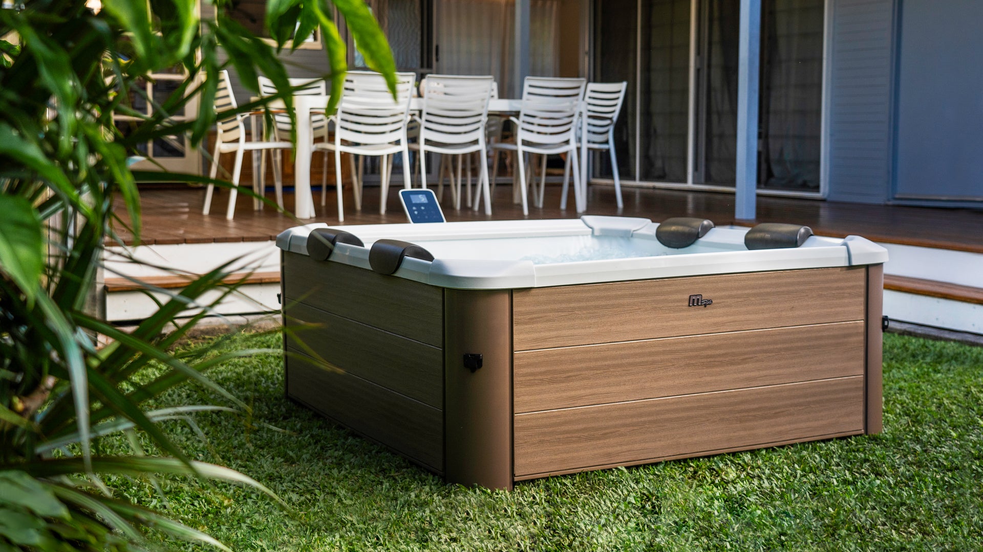 Portable square hot tub jacuzzi spa with brown wood-like exterior in backyard area with outdoor furniture on a deck in the background