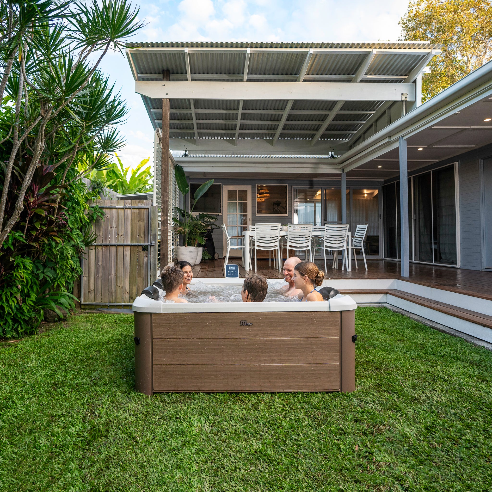 Happy family of five enjoying a spa in a square framed hot tub jacuzzi with wood like exterior in a backyard with a modern house and outdoor furniture.